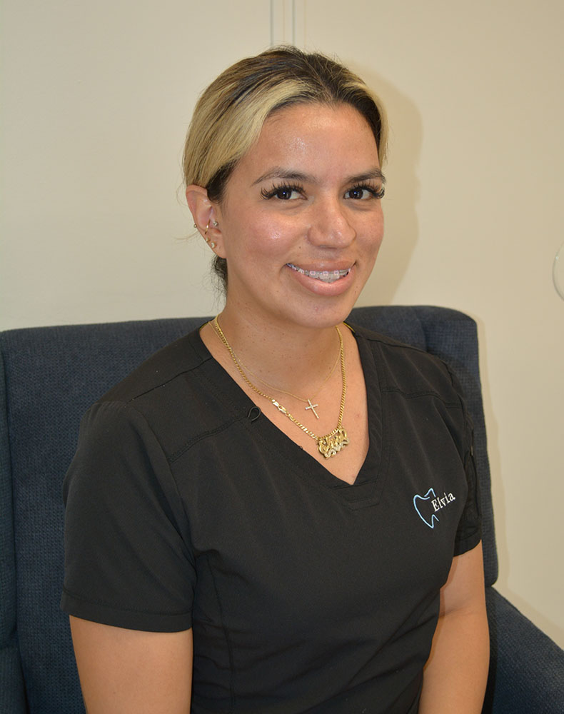 The image shows a woman sitting on a chair with a professional attire, likely a nurse or healthcare worker, wearing a stethoscope around her neck and a name tag. She has a friendly expression and appears to be in an office setting.