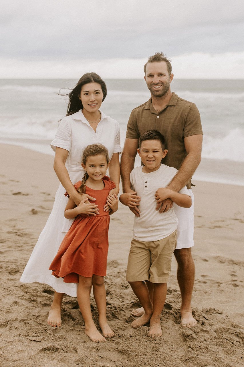 The image shows a family of four people standing on sandy beach, with a man and woman standing closer together than two children who are positioned slightly apart from them  all are smiling and appear to be posing for the photograph.
