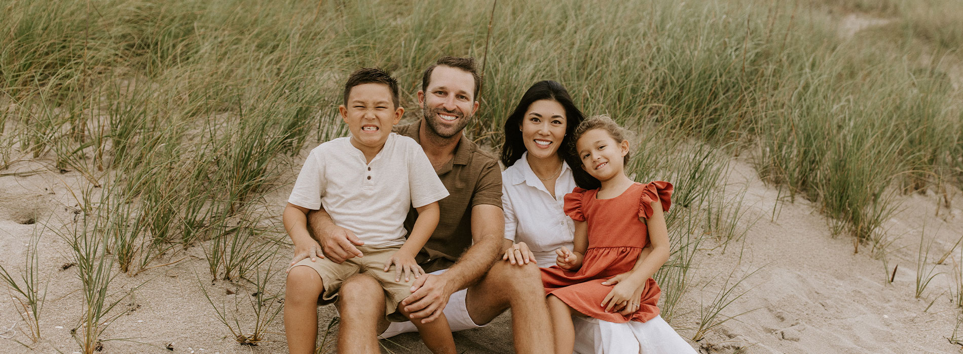 A family of four posing on a sandy beach with dunes in the background.