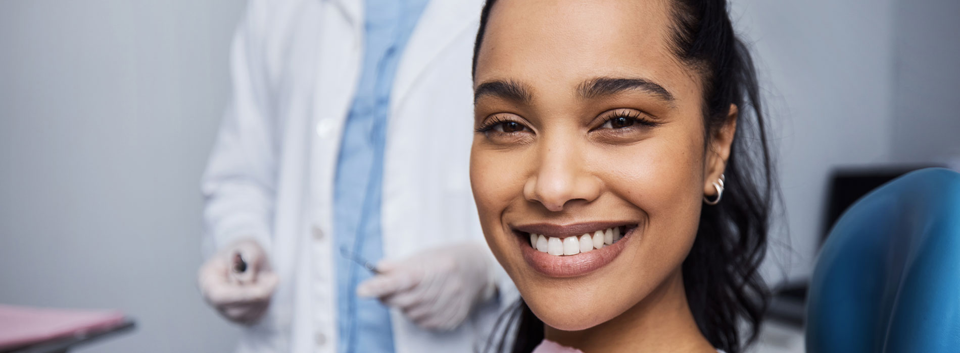 A person is seated in a dental chair, receiving care from a dental professional who stands behind them.