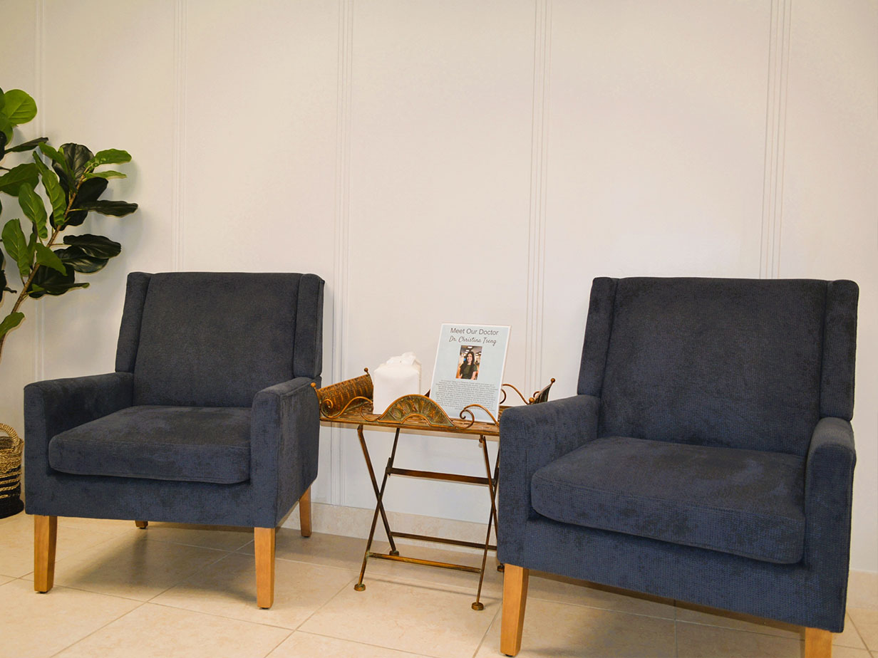 The image shows two blue armchairs placed next to each other against a white wall with a plant on the left side, and there s a small table between them.