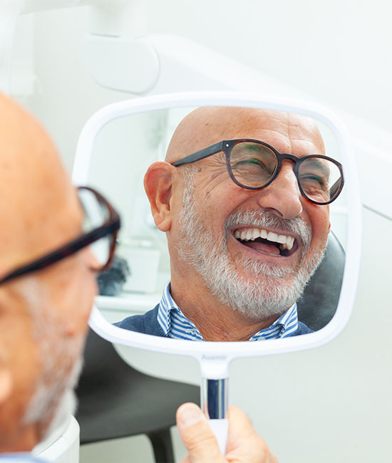 The image shows a man with glasses smiling at his reflection in a mirror while sitting in a dental chair, with a dental professional s hand visible holding the mirror.