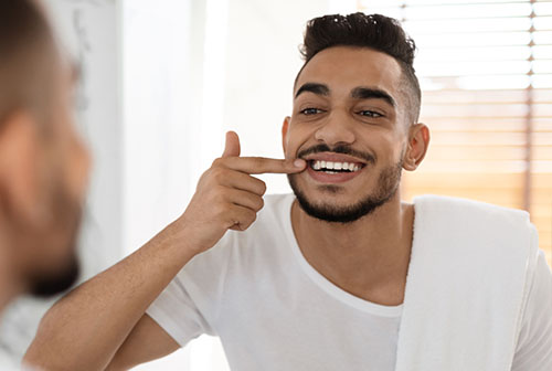 The image shows a man with a beard, smiling broadly while looking at his own reflection in a mirror, holding his nose with one hand. He appears to be in a bathroom setting, wearing a white towel around his neck and has a playful expression on his face.