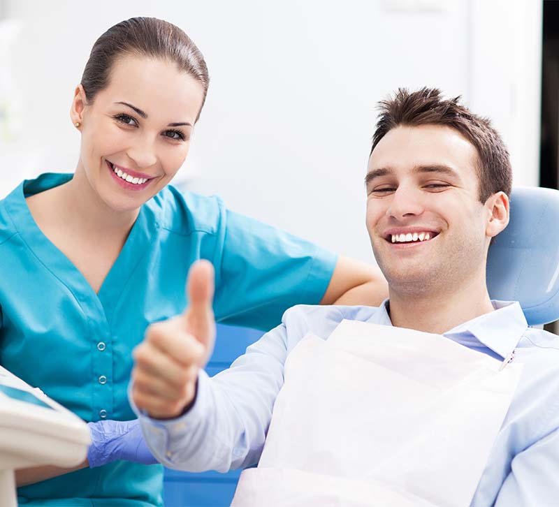 The image shows a dental professional, presumably a dentist, giving a thumbs-up while sitting at a desk with medical equipment, with a patient seated in a chair receiving dental care.